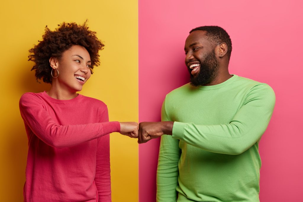 Picture of a woman on pink shirt in a yellow background chop knucking a man on green shirt in a pink background.