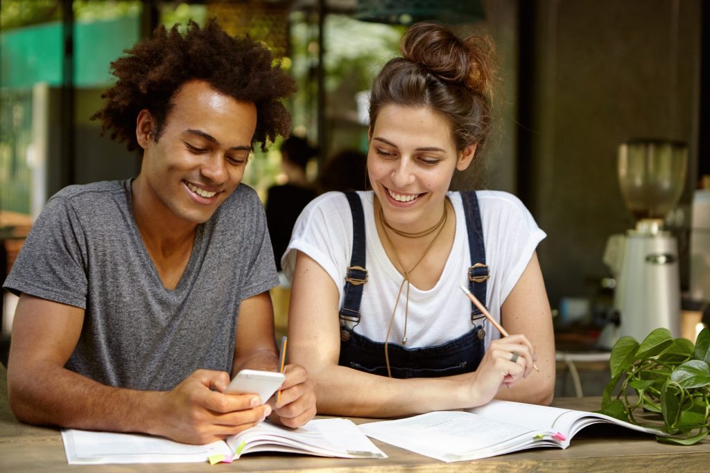 A man on grey shirt holding his phone and smiling sitting next to a woman wearing a white shirt under jean overalls smiling as they both look into his phone.