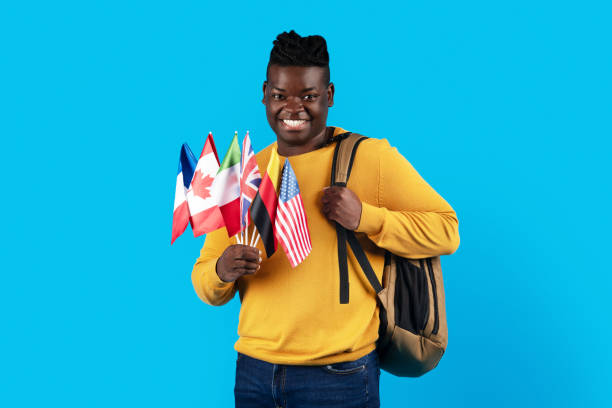 Happy black man holding stack of international flags and backpack, smiling, standing over blue background.